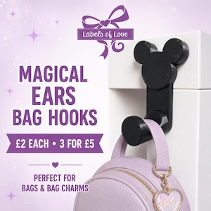 May include: A black bag hook shaped like a mouse head silhouette with rounded ears, mounted on a white surface. A light purple circular bag with a gold zipper and a heart-shaped charm hangs from the hook. Text reads Labels of Love, MAGICAL EARS BAG HOOKS, £2 EACH 3 FOR £5, PERFECT FOR BAGS & BAG CHARMS.