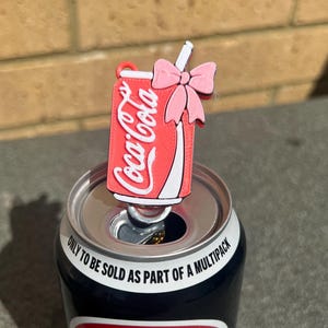 May include: A red and white Coca-Cola can-shaped charm with a pink bow and straw detail, placed on top of a Pepsi can. The Coca-Cola charm features the Coca-Cola logo. The Pepsi can has a red and white stripe.