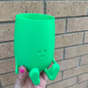 May include: A vibrant green, 3D-printed cup featuring a smiling face, arms, and feet. The cup is held in a hand. The cup has a cylindrical shape with a rounded base and is set against a brick wall.