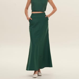 May include: A woman wearing a dark green two-piece outfit. The top is a sleeveless wrap-style crop top with a V-neck. The matching skirt is a floor-length, high-waisted design with pockets. The outfit is made of a solid, smooth fabric.