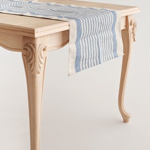 Op de afbeelding: Een lichtgekleurde houten tafel met sierlijke poten, gedrapeerd met een blauw-wit gestreepte linnen tafelloper. De loper hangt over de rand van de tafel. De tafel staat tegen een witte achtergrond.