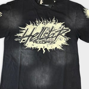May include: A black short-sleeved t-shirt with a faded wash effect. The centre features a cream-coloured graphic with flame-like edges and the text Hellstar RECORDS. Additional flame and text graphics appear on both sleeves, with a tag at the collar.
