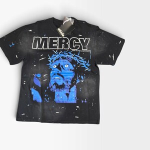 Könnte beinhalten: Ein schwarzes Kurzarm-T-Shirt mit einer blauen Grafik eines Gesichts, das eine Dornenkrone trägt. Das Wort MERCY ist in großen weißen Buchstaben über der Grafik gedruckt. Das T-Shirt hat einen weißen Farbspritzer-Effekt und ein schwarzes Markenetikett.