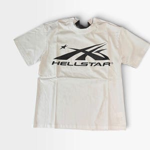 Könnte beinhalten: Ein weißes Kurzarm-T-Shirt mit einer schwarzen Grafik auf der Brust. Die Grafik zeigt einen stilisierten Stern und geschwungene Linien über dem Text HELLSTAR in einer fetten serifenlosen Schrift. Das T-Shirt hat einen Rundhalsausschnitt.