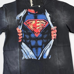 May include: A black short-sleeve t-shirt featuring a graphic print of a blue textured chest piece with a red diamond-shaped emblem. The design shows a shirt being ripped open to reveal the emblem, which includes a star and a profile silhouette.