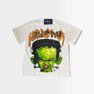 Peut inclure: Un t-shirt blanc à manches courtes avec une impression graphique représentant un personnage à la peau verte, aux cheveux foncés et portant un boulon métallique au cou. Au-dessus, le mot HELLSTAR est imprimé en orange avec des contours noirs.