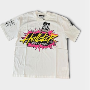 Könnte beinhalten: Ein weißes Kurzarm-T-Shirt mit einer zentralen Grafik, die den gelben Schriftzug Hellstar und den weißen Schriftzug RECORDS über einer pinkfarbenen, flammenartigen Form zeigt. Ein schwarzes Etikett mit weißem Logo und HELLSTAR am Kragen.