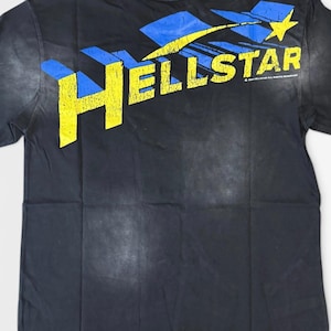 Könnte beinhalten: Ein schwarzes Kurzarm-T-Shirt mit einem verwaschenen gelb-blauen Grafikdruck auf der oberen Brustpartie. Das Design zeigt den Schriftzug HELLSTAR in großen gelben Blockbuchstaben sowie einen gelben Stern und blaue geometrische Formen darüber.