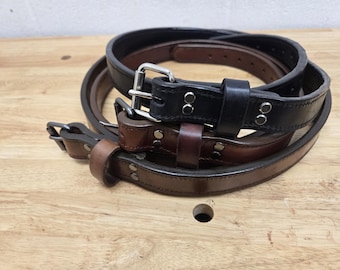 May include: A stack of leather belts in various shades of brown and black. Each belt features a metal buckle and is secured with rivets. The belts are arranged on a wooden surface, showcasing their different colors and styles.