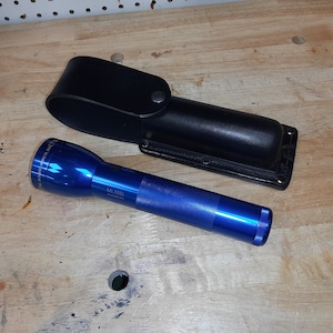May include: A blue metal flashlight with the brand name "Maglite" printed on the side. The flashlight is next to a black leather holster.