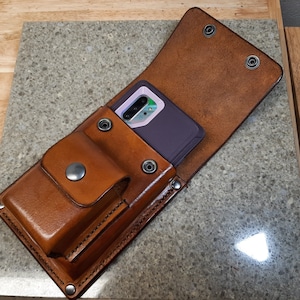 May include: A brown leather phone case with a flap that folds over to protect the phone. The case has a separate pocket for carrying other items. A purple phone is inside the case.