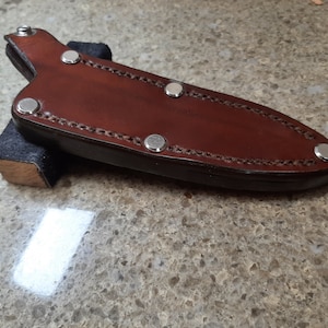 Puede incluir: Una funda de cuchillo de cuero marrón con remaches plateados. La funda está doblada y tiene un borde cosido.