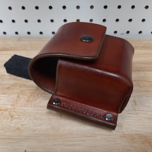 May include: A brown leather pouch with a flap closure and a metal snap. The pouch has a black strap and is secured with metal rivets.