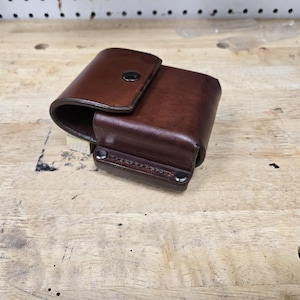 May include: A brown leather pouch with a flap secured by a snap. The pouch has a rectangular shape and is stitched along the bottom. The leather has a rich, dark color and a slightly worn appearance.