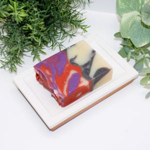 May include: A colorful handmade soap bar with red, purple, black, and cream swirls, resting on a white rectangular soap dish with a brown rim. The soap has a marbled appearance. The background includes green foliage.