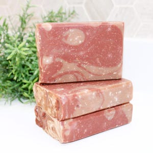May include: Three rectangular bars of handmade soap are stacked. The soap has a marbled appearance with shades of red and tan. The top bar has a large, light-colored swirl. The soap is a natural product.