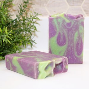 May include: Two rectangular bars of handmade soap. The soap has a swirl design of purple and green colors. The soap is sitting on a white surface with a green plant in the background.