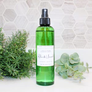 May include: A green glass spray bottle with a black nozzle, labeled "Relax & Unwind" and "Linen & Room Scented Spray." The bottle contains a clear liquid and is 6 oz / 227 g. The background includes greenery and a white surface.