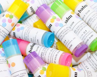 May include: A collection of colorful lip balm tubes with various colored caps, including yellow, blue, pink, green, and purple. Each tube has a white label with the word "balm" and other text. The lip balms are 0.2 oz (6 gr).