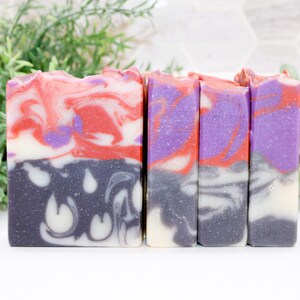 May include: Four bars of handmade soap with a layered design. The top layer features red, white, and purple swirls, while the bottom layer is dark gray with a white pattern. The soaps are arranged in a row, with green foliage in the background.