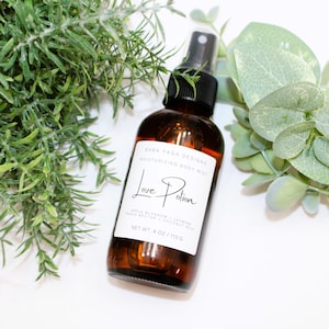 May include: A brown glass bottle of moisturizing body mist with a black spray nozzle. The label reads "Love Potion" and lists ingredients. The bottle is 4 oz / 113 g. Green foliage surrounds the bottle.