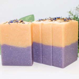 May include: Two-tone soap bars with a lavender base and a peach-colored top, adorned with dried flower petals. The soap bars are rectangular and have a smooth texture. The image showcases the soap bars with a white background and greenery.