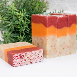 May include: Handmade soap bars in a natural theme. The soaps are layered with colors of red, orange, and beige. One bar is decorated with red and gold glitter. The soaps are rectangular and stand upright.