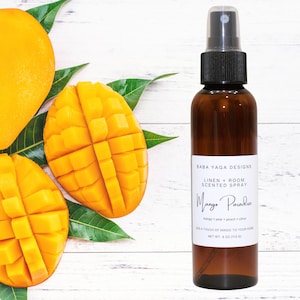 May include: A brown glass spray bottle with a black cap, labeled "Mango Paradise" with text indicating it is a linen and room scented spray. The image includes fresh mango slices and green leaves, with a net weight of 4 oz (113 g).