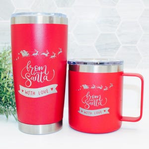 May include: Two red stainless steel tumblers with a white design that says "from Santa with love". The larger tumbler is taller and has a lid. The smaller tumbler has a handle and is shorter. Both tumblers have a silver band around the bottom.