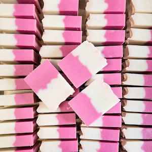 May include: Rows of handmade soap bars with a two-tone design. The bars are white and pink, with a wavy line separating the colors. The soap bars are stacked neatly, with a few bars scattered on top.
