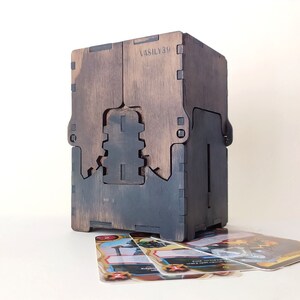 May include: A wooden box with a dark brown stain and a black interior. The box has a slot on the front for holding cards. The box is sitting on a white surface with several playing cards in front of it.