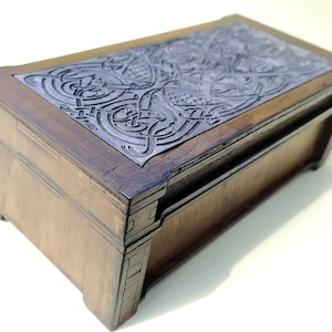 May include: A wooden box with a hinged lid. The lid is decorated with an intricate silver-colored Celtic knot design.