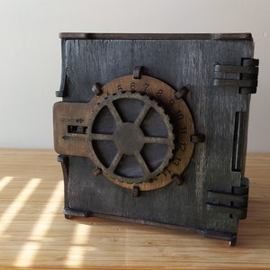 May include: A wooden box shaped like a safe with a metal dial and a handle. The dial has numbers 1 through 13 and the handle has a lever that can be moved up and down. The box is painted a dark grey colour and has a distressed look.