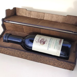 Puede incluir: Una caja de madera con una botella de vino tinto dentro. La caja tiene una tapa que se levanta y está decorada con un diseño intrincado. La botella está etiquetada con "Habich", "Muller Thurgau", "Dry Red Wine" y "Dyrrat El-Hocq".