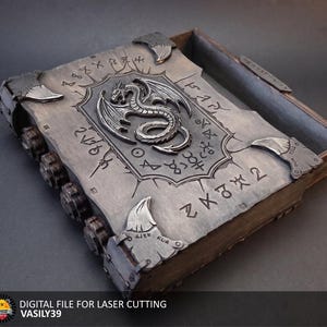 May include: A rectangular wooden box with a sliding lid featuring a silver-toned dragon emblem in the center. The lid is engraved with runic symbols and has metallic corner accents. The side of the box displays four circular gear-like components.
