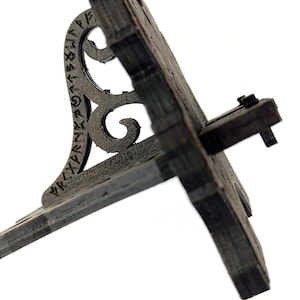 May include: A weathered, dark gray wooden stand with a decorative, scrolled support. The stand features carved runes and a hinged, angled surface. The design suggests a historical or fantasy theme.