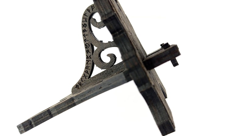 May include: A weathered, dark gray wooden stand with a decorative, scrolled support. The stand features carved runes and a hinged, angled surface. The design suggests a historical or fantasy theme.