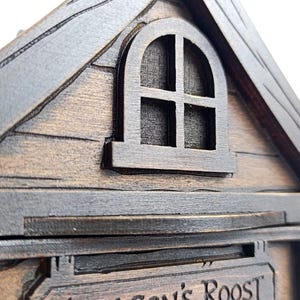 May include: A dark brown wooden structure shaped like a house with a gabled roof and a small arched window featuring four panes. The exterior displays wood grain textures and carved lines. A sign below the window reads Dragon's Roost.