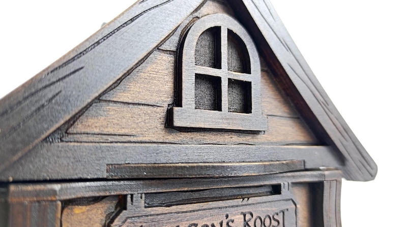 May include: A dark brown wooden structure shaped like a house with a gabled roof and a small arched window featuring four panes. The exterior displays wood grain textures and carved lines. A sign below the window reads Dragon's Roost.