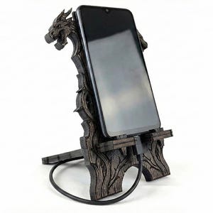 May include: A dark brown wooden phone stand with a dragon head design. The stand holds a black smartphone, with a charging cable connected. The stand has a detailed, carved appearance and is set against a white background.