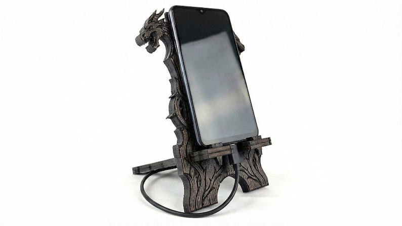 May include: A dark brown wooden phone stand with a dragon head design. The stand holds a black smartphone, with a charging cable connected. The stand has a detailed, carved appearance and is set against a white background.