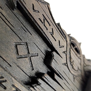May include: Close-up of a dark wooden object with carved runes and geometric shapes. The wood has a distressed appearance with visible grain and a weathered texture. The design suggests a historical or fantasy theme.