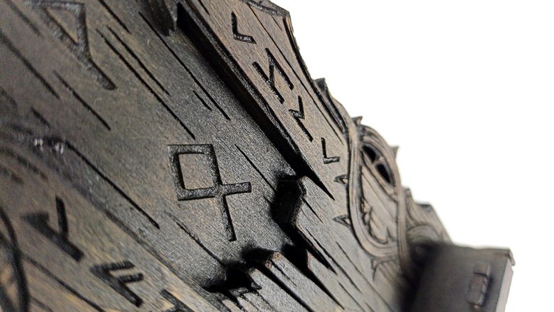 May include: Close-up of a dark wooden object with carved runes and geometric shapes. The wood has a distressed appearance with visible grain and a weathered texture. The design suggests a historical or fantasy theme.