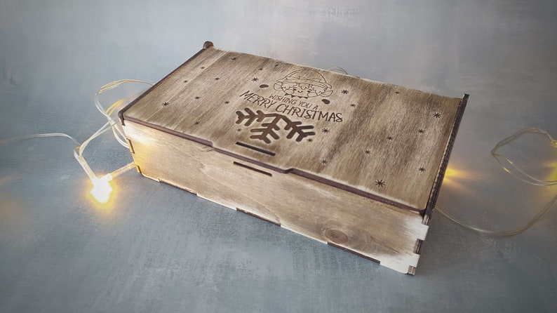 May include: A wooden box with a hinged lid. The lid is decorated with a laser-cut design of a snowflake and the text "Wishing you a Merry Christmas".