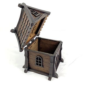 May include: A dark brown wooden box shaped like a small house with an open hinged roof. The structure features textured walls, a window with a grid frame, and a roof with a scalloped shingle pattern. The interior is hollow and square-shaped.