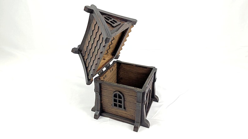 May include: A dark brown wooden box shaped like a small house with an open hinged roof. The structure features textured walls, a window with a grid frame, and a roof with a scalloped shingle pattern. The interior is hollow and square-shaped.