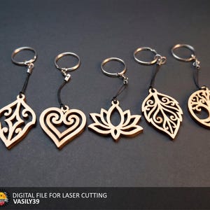 Può includere: Cinque portachiavi in legno chiaro con intricati disegni tagliati al laser, tra cui una forma a diamante, un cuore, un fiore di loto, una foglia e un albero della vita. Ogni pezzo è fissato a un cordino nero e a un anello in metallo argentato. Testo: DIGITAL FILE FOR LASER CUTTING VASILY39.