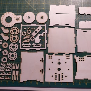 May include: A set of laser-cut wooden pieces for building a model. The pieces are arranged on a green cutting mat. The pieces include a variety of shapes, including circles, squares, and rectangles. There are also some smaller pieces that appear to be gears.
