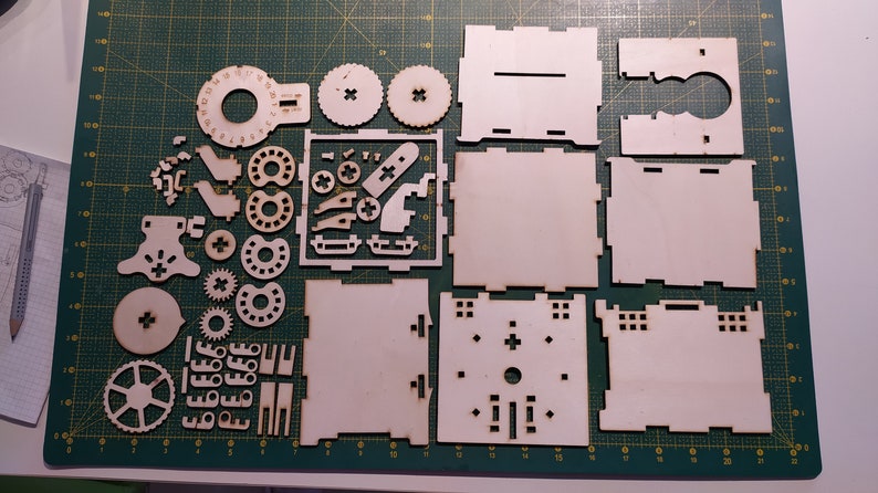 May include: A set of laser-cut wooden pieces for building a model. The pieces are arranged on a green cutting mat. The pieces include a variety of shapes, including circles, squares, and rectangles. There are also some smaller pieces that appear to be gears.