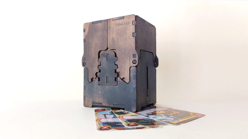 May include: A wooden box with a dark brown stain and a black interior. The box has a slot on the front for holding cards. The box is sitting on a white surface with several playing cards in front of it.